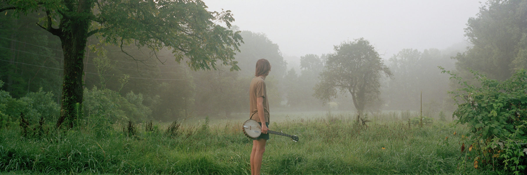 white man stands with alone in a hazy field dotted with trees. He holds a banjo and has his back turned toward the camera.