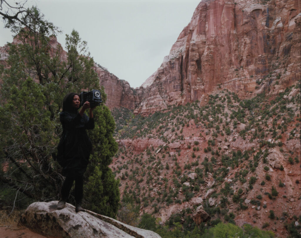 A woman with dark skin stands in front of red cliffs dotted with scrubby brush. She holds a large format camera in front of her face.